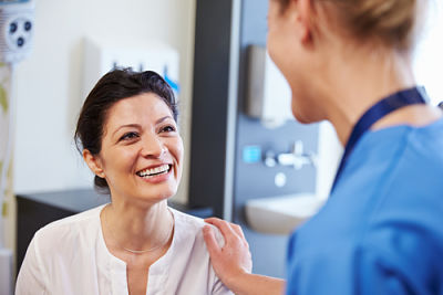 Female Nurse is smiling at a female patient who is sitting down in front of her smiling.