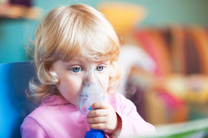 Female toddler is holding a breathing mask up to her face during respiratory therapy.
